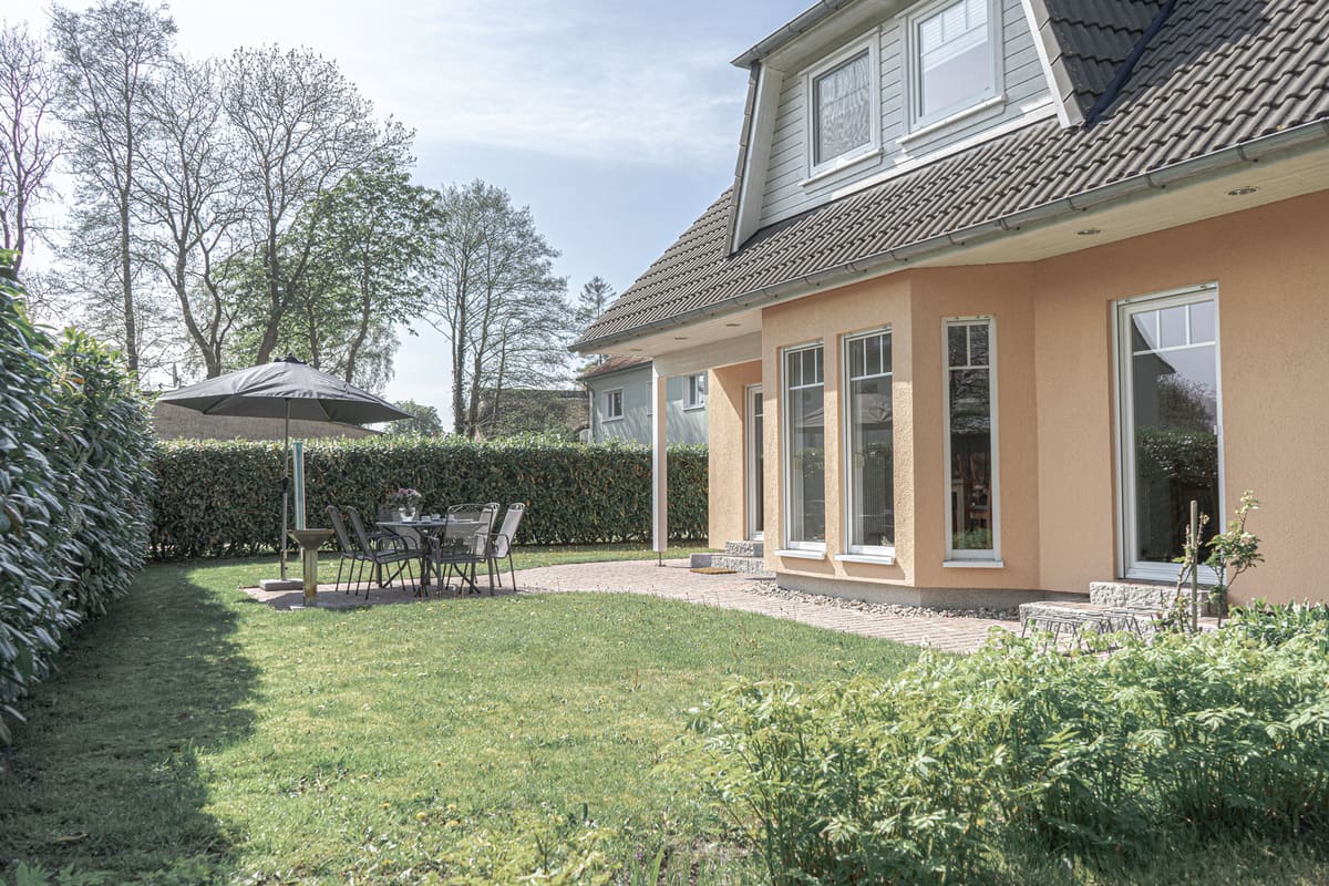 Terrasse mit Gartentisch, Stühlen und Sonnenschirm, geschützt von einer hohen Hecke