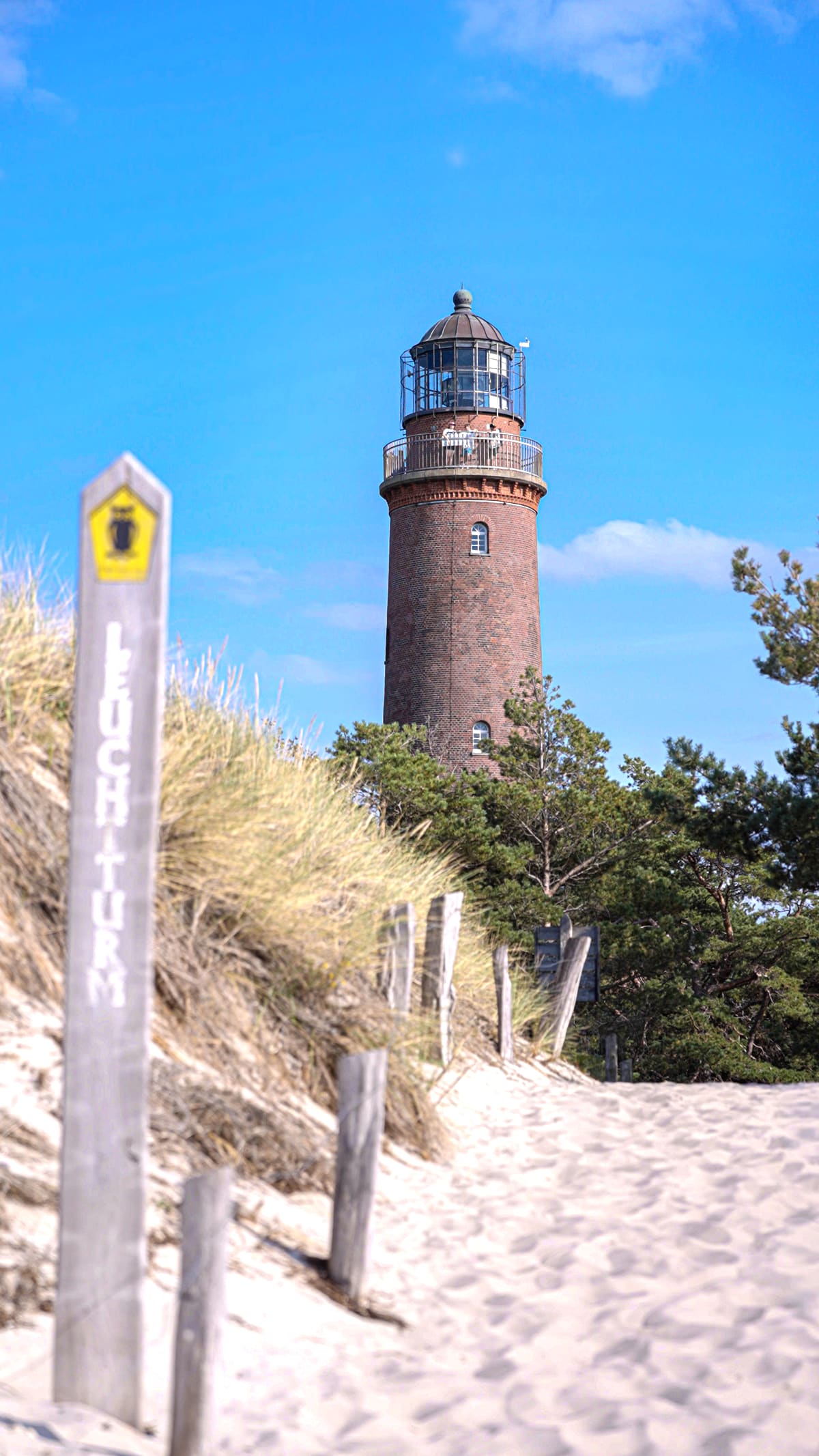 Hölzerner Wegweiser „Leuchtturm“ im Dünensand, dahinter der Backsteinturm vor kräftig blauem Himmel