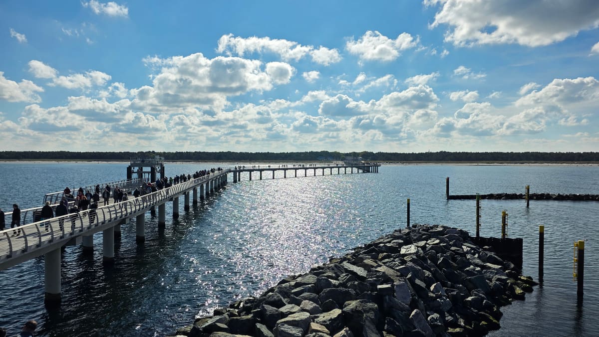Seebrücke mit Spaziergängern führt über das Wasser hinaus, rechts eine Steinmole, die Sonne spiegelt sich in der Ostsee